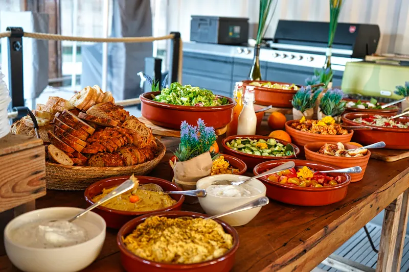 Buffet mit Brot, Salaten und Dips in Terrakotta-Schalen, im Hintergrund der Grill